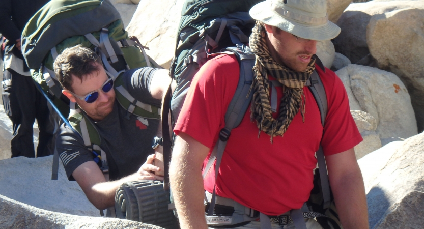 One backpacker helps adjust the backpack of another near large boulders. 